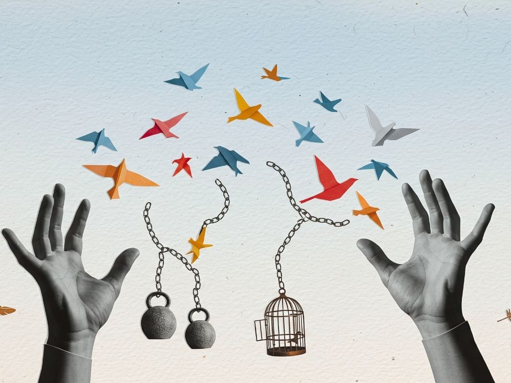 A pair of raised up arms letting go of broken chain that has been holding them back while a cage opens, weights drop and uncaged birds fly free.