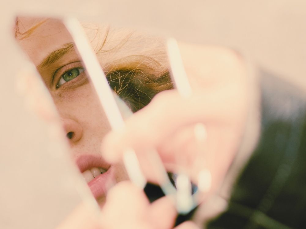 A woman looking at herself in a broken mirror pieces depicting the challenges of seeing oneself clearly and recognizing toxic stress and trauma injuries.