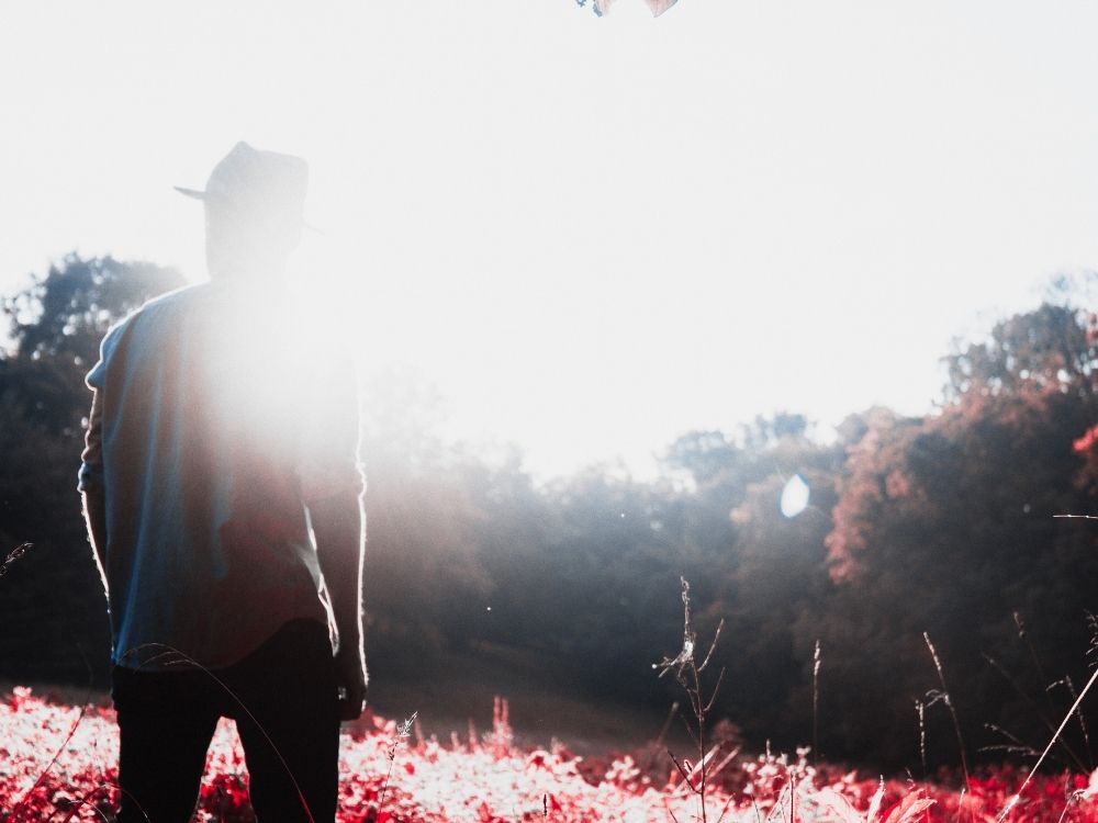 112 A man wearing a hat walking into a treed area with beautiful sunlight landing all around.